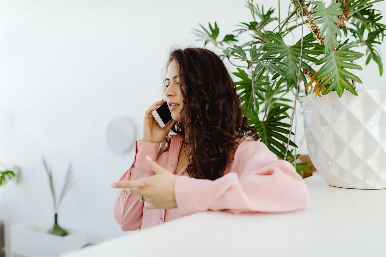 A woman on a phone call at a bright desk with plants