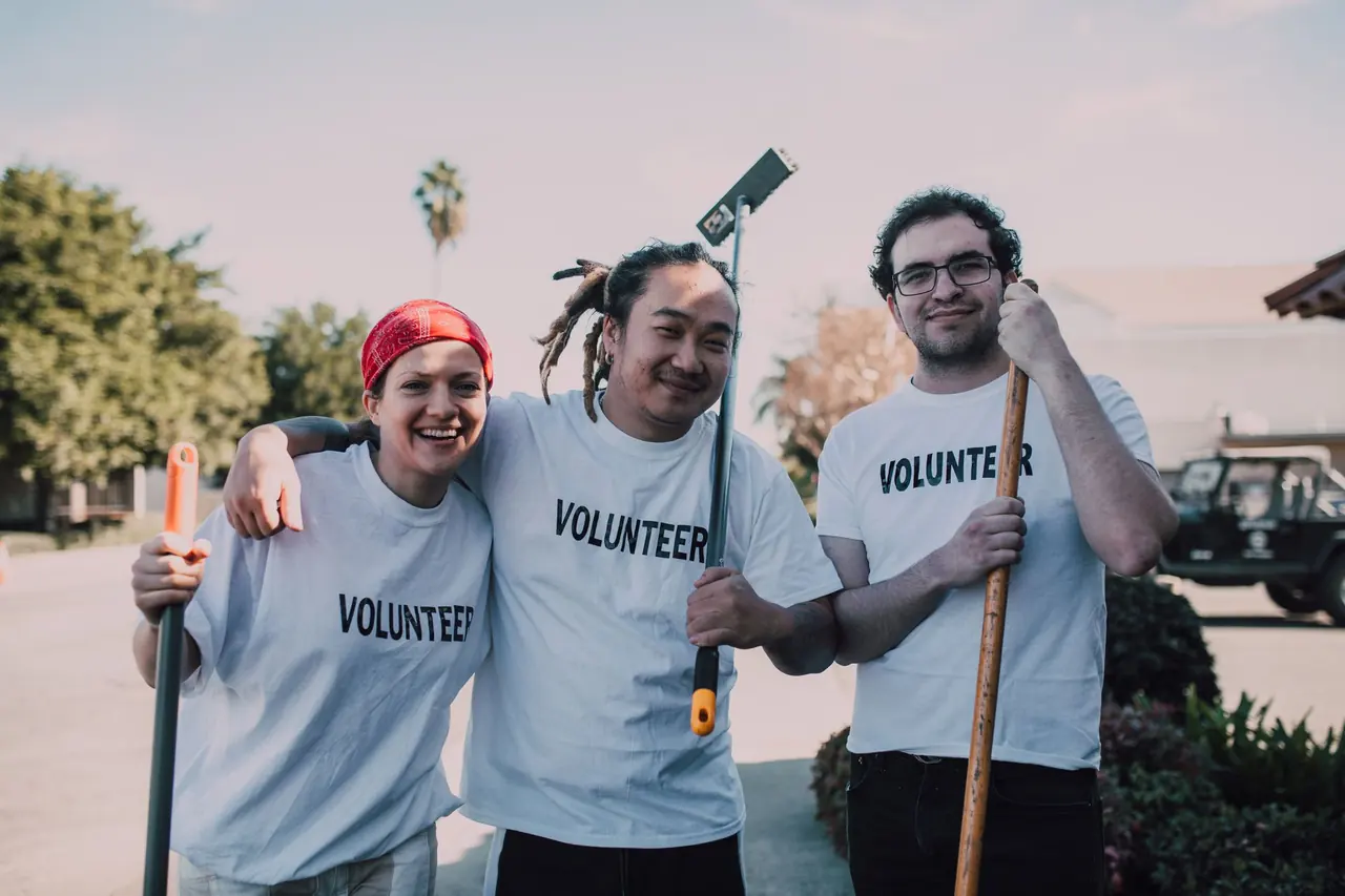 Three smiling volunteers holding rakes and brooms at a community cleanup